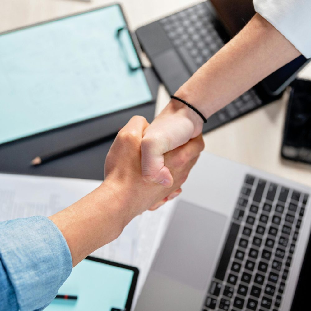 Two people shaking hands over laptops and documents in an office, symbolizing a business deal.